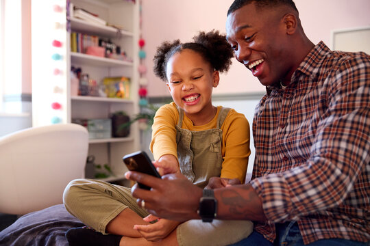 Father And Daughter At Home In Girl's Bedroom Laughing At Pictures On Mobile Phone