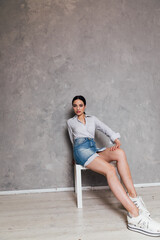 a fashionable woman in a denim skirt sits on a chair with a gray background