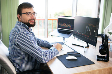 Fototapeta na wymiar Side view a man looks into the camera smiling with white teeth analyzing the graphs on the monitors in the office. Concept: broker, cryptocurrency, indexes, trading online. investment, data on stock