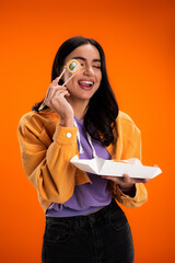 Positive woman holding takeaway sushi and chopsticks while sticking out tongue isolated on orange.