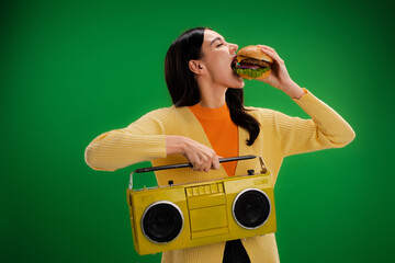 young and hungry woman with boombox eating tasty burger isolated on green.