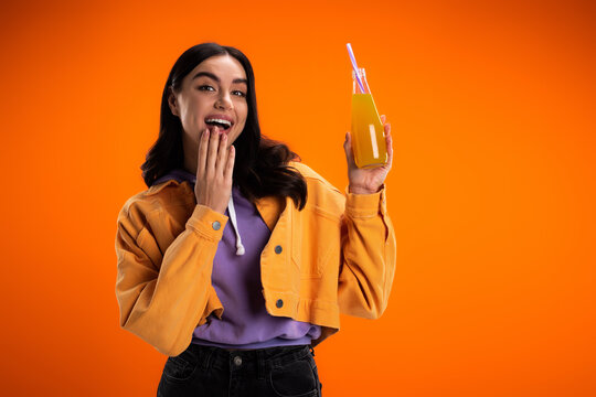 Excited Woman Looking At Camera While Holding Bottle With Juice Isolated On Orange.