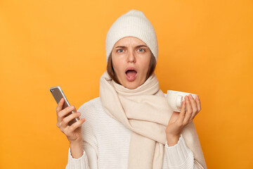 Shocked amazed astonished woman wearing warm jumper, hat and scarf posing isolated over yellow background, standing with cup of beverage and smart phone, looking at camera with big eyes and open mouth