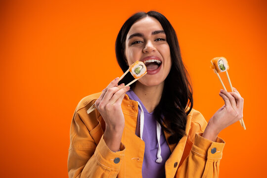 pleased woman in trendy jacket eating tasty sushi roll and looking at camera isolated on orange.