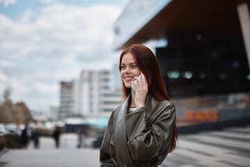 Portrait of a beautiful young woman in the city with a phone smile with teeth with red flying hair in fashionable clothes in the city background, lifestyle in the city