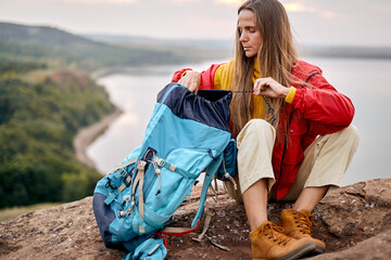 Confident caucasian female traveller unpacking backpack, river in the background, travelling alone,...
