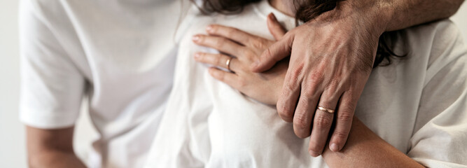 Fototapeta na wymiar Human hands with wedding rings close-up young heterosexual couple gentle embrace wearing white t-shirts.