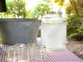 Transparent glass jar with tap filled by fresh homemade lemonade placed on table near glasses and metal bucket against trees