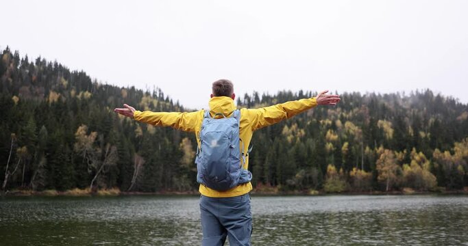 Happy male traveler standing with arms wide apart in front of mountain lake back view 4k movie slow motion