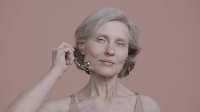 CU Portrait of very beautiful grey-haired 60s mature senior adult female using cosmetics face roller. Studio shot, soft lighting. No make up, clean skin