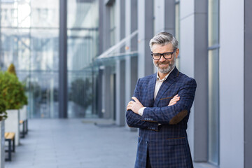 Portrait of mature gray-haired businessman, senior boss smiling and looking at camera with arms...