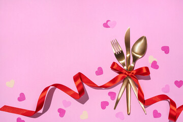 Golden fork, knife, spoon tied with red ribbon on pink background. Valentine's Day or festive romantic dinner.