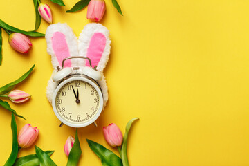 Alarm clock in rabbit ears surrounded by tulips. Easter spring change of seasons.