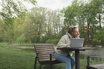 Fototapeta premium Woman working out of office in the park near the river. Concept of remote work at any time and place 