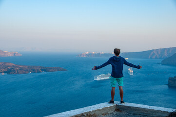 A man has his arms lifted on the Cliffs of Santorini during Sunrise as cruise ships go by in the beautiful Bay below