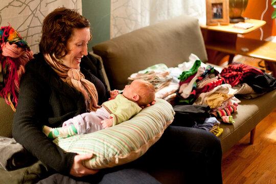 A Smiling Mother With Red Hair Plays With Her Baby On A Couch Amid Piles Of Messy Laundry At Night In A Home.