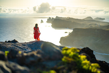 Fototapeta premium Woman in red dress enjoys panoramic view from steep cliff over seascape and along rugged foothills of Madeira coast at sunrise. Ponta do Bode, Madeira Island, Portugal, Europe.