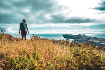 Fototapeta premium Backpacker woman enjoys hike along a steep cliff overlooking the sea and the rugged foothills of Madeira's coast in the morning. Ponta do Bode, Madeira Island, Portugal, Europe.