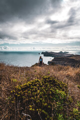 Backpacker woman enjoys hike along a steep cliff overlooking the sea and the rugged foothills of Madeira's coast in the morning. Ponta do Bode, Madeira Island, Portugal, Europe.