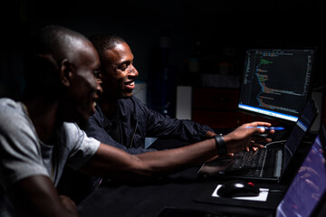 Two black males smiling while looking at code in a dark room