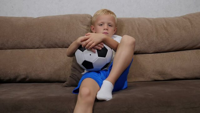 A small six-year-old boy is sitting on the sofa at home and holding a soccer ball.
