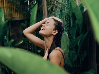 A woman takes a shower and washes her head and hair outdoors, closed eyes and a smile on the background of tropical plants, palm trees, green banana leaves, summer rain