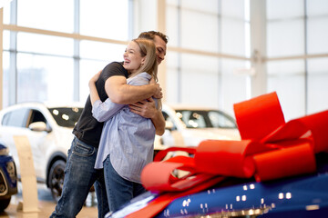 Guy Shows New Car To Girlfriend. Present Concept. Hugging. Automobile Salon. Make A Decision. Gift Ribbon. Good Offer. Happy Together. Successful Buying. man makes a gift car to wife. in good mood.