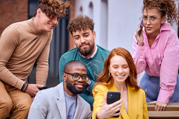 diverse men and women colleagues take photo on smartphone outdoors, smiling, spending time together during break time. african and caucasian males and females in stylish formal clothes sit on bench