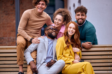 Portrait of successful group of people enjoying free time together outdoors. Men and women in stylish formal outfits after working in ecommerce business office, laughing,sitting on bench