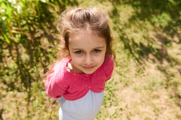 Close-up cute little girl in blue summer dress and pink bolero, smiles looking at camera, standing in nature background