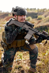 Fototapeta premium Brave caucasian male in combat uniform is standing in nature alone waiting for operation, strong soldier man in combat uniform holding rifle weapon in hands, posing, leaned on one knee