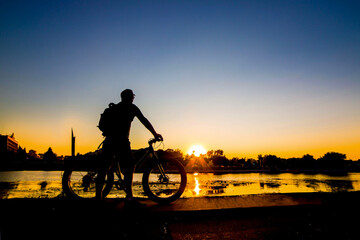 silhouette of a cyclist with a fatbike standing on the river bank at sunset