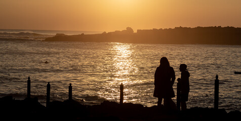 silhouettes of mother and child enjoying the sunset at the sea in winter, with silhouette of man returning from surfing