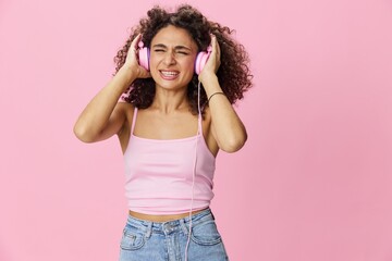 Happy woman wearing headphones with curly hair listening to music and singing along with her eyes closed in a pink T-shirt and jeans on a pink background, copy space