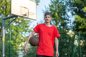 Cute young teenager in red t shirt with a ball plays basketball on court. Teenager dribbling the ball,  running in the stadium. Sports, hobby, active lifestyle for boys	