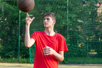 Cute young teenager in red t shirt with a ball plays basketball on court. Teenager dribbling the ball,  running in the stadium. Sports, hobby, active lifestyle for boys	