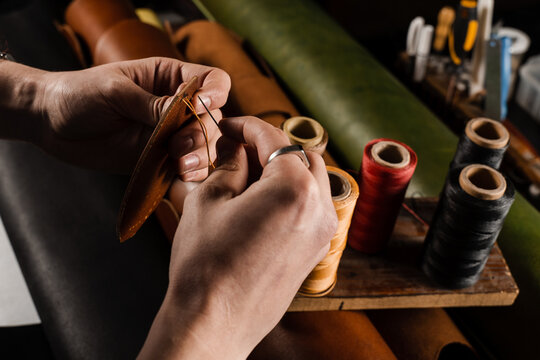 Process of stitching genuine leather. Craftsman sews genuine leather using needle and thread for creation natural leather products. Equipment for genuine leather production in workshop.