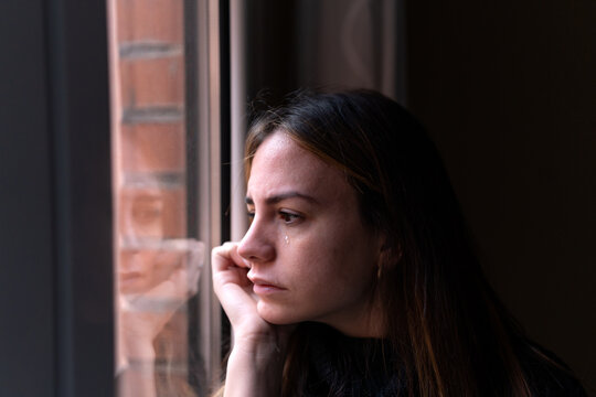 Side view of lonely lady with dark hair looking out of window and touching face while crying
