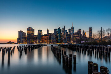 Fototapeta premium New York, USA - April 25, 2022: Long exposure of the Lower Manhattan skyline at sunset with an old Brooklyn pier in the foreground