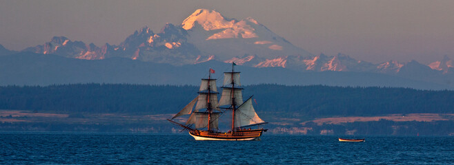 Nineteenth century replica ship, Hawaiian Chieftain, sets sail in Penn Cove, Washington, USA at sunset with Mount Baker in the distance.