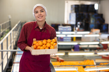 Happy young female worker of citrus sorting factory showing ripe selected mandarin oranges packed...