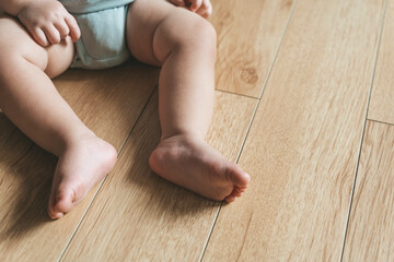 Baby sits on a wooden laminate floor. Bare legs and feet of a baby in the living room floor