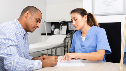 Obraz premium Qualified Latin American female beautician sitting at table with man client in aesthetic cosmetology clinic, filling medical card..