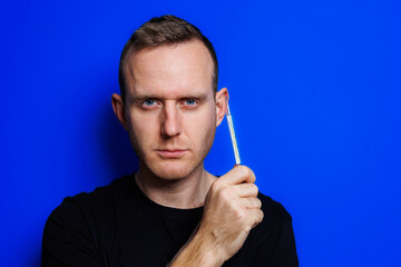 A young man in a black T-shirt on a blue background stands with a thermometer in his hands. An increase in body temperature, feeling sick. Weakened immunity. Selective focus