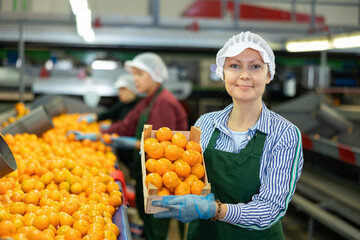 Portrait of smiling woman professional sorter standing with box of selected tangerines at fruit processing factory © JackF