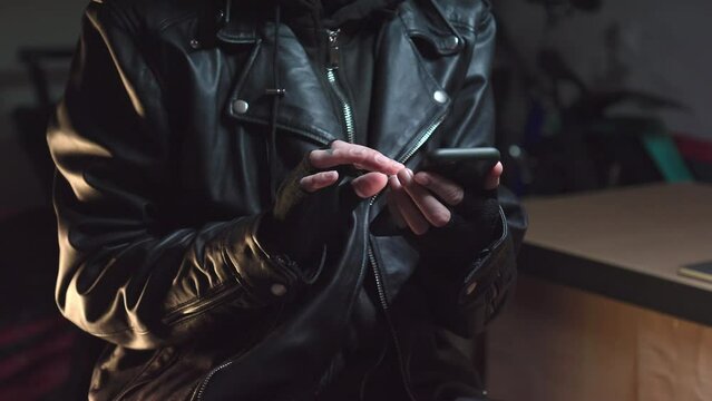 Hacker wearing mask using phone dressed in leather jacker and gloves.