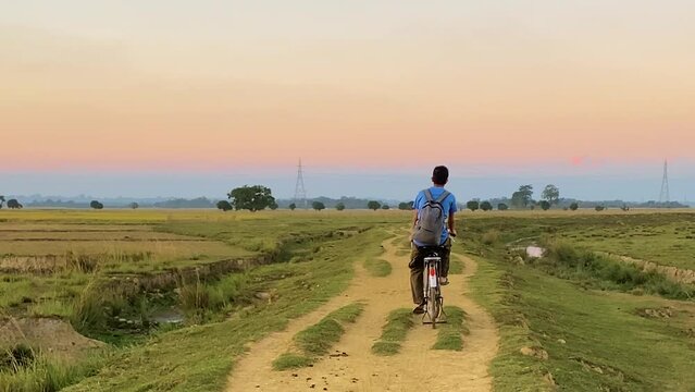 Young village boy with backpack cycle in rural path through agriculture fields in Bangladesh, back view