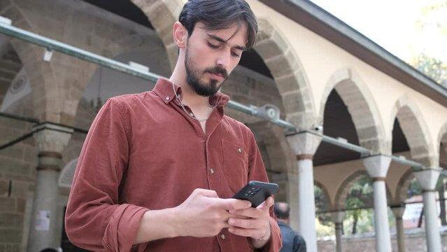 A bearded man is using his phone in front of the mosque, a young person is texting with a smartphone, a mosque is a place of worship for Muslims