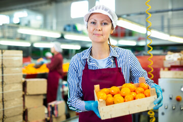 Portrait of smiling woman professional sorter standing with box of selected tangerines at fruit processing factory © JackF