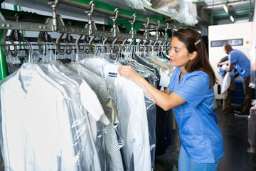 Positive female dry-cleaning worker preparing garments for collection, hanging clean clothes packed in plastic bags on rail